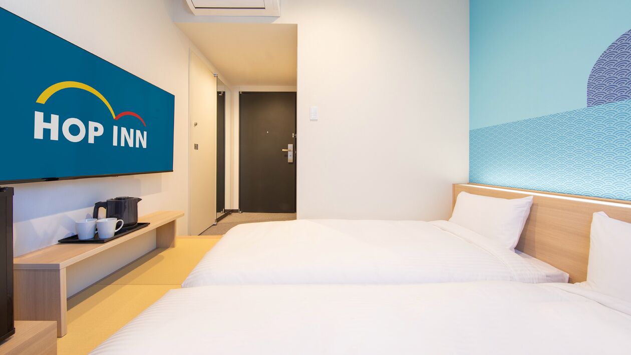 Quarto de hotel com cama, televisão fixada na parede, cafeteira e porta que dá para outro aposento.