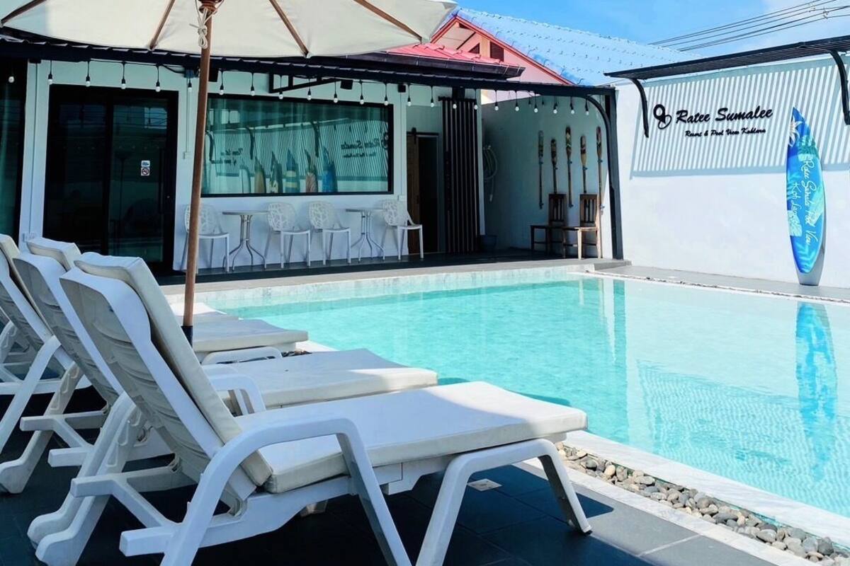 A pool area with white lounge chairs and a large umbrella, a restaurant with outdoor seating, and a surfboard on the wall.