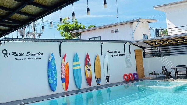 Una piscina con tablas de surf de colores en la pared, un área cubierta con luces colgantes y un letrero que indica Resort & Pool Ratee Sumalee.