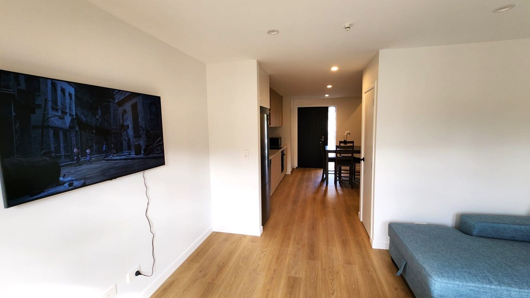 Un appartement moderne doté d’un plancher en bois, d’un téléviseur à écran plat fixé au mur, d’un coin repas et d’un canapé bleu.