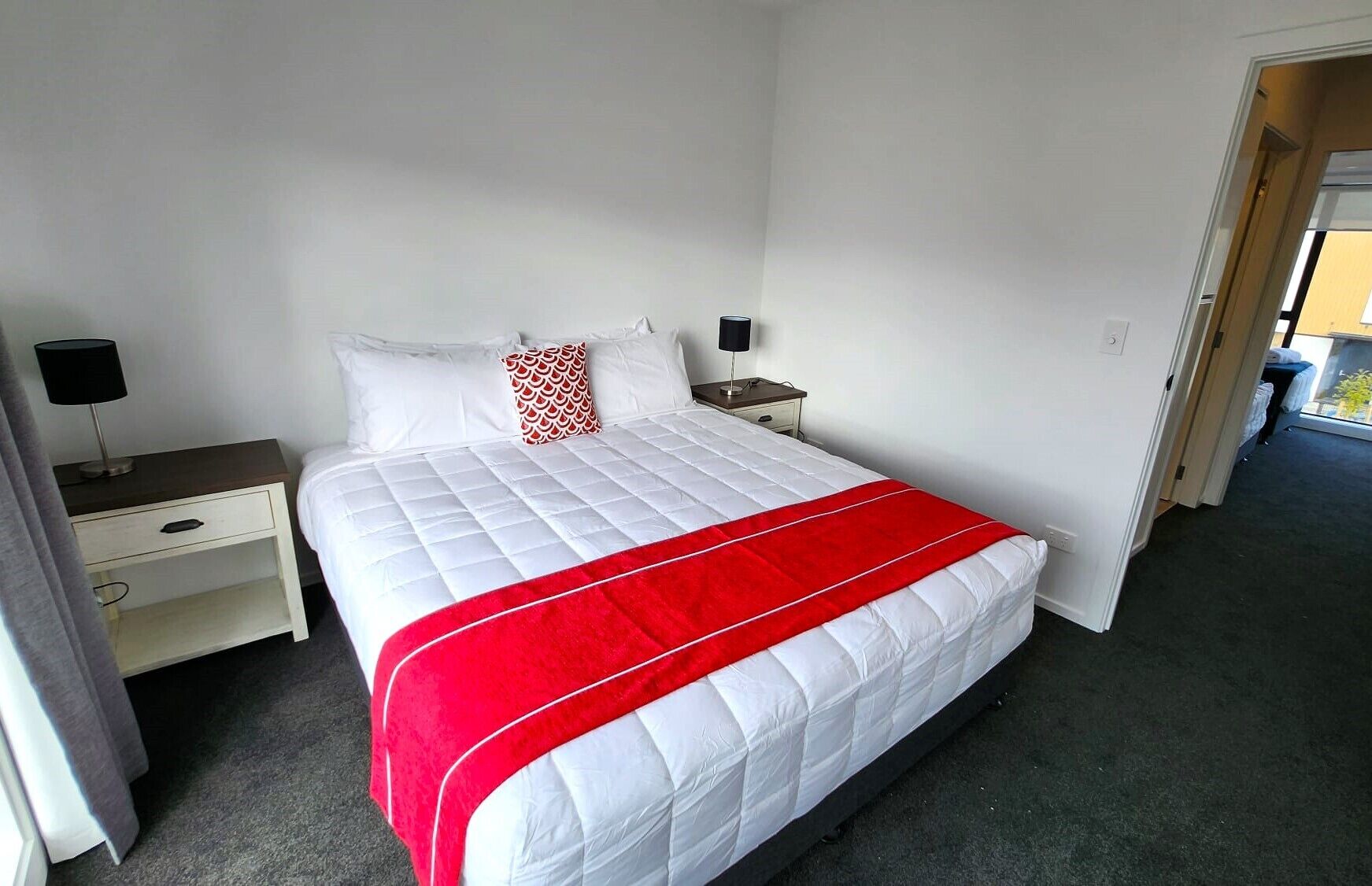 A hotel room with a bed, two bedside tables, a nightstand, a red and white bedspread, and a white pillow with a red pattern.
