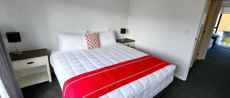 A hotel room with a bed, two bedside tables, a nightstand, a red and white bedspread, and a white pillow with a red pattern.