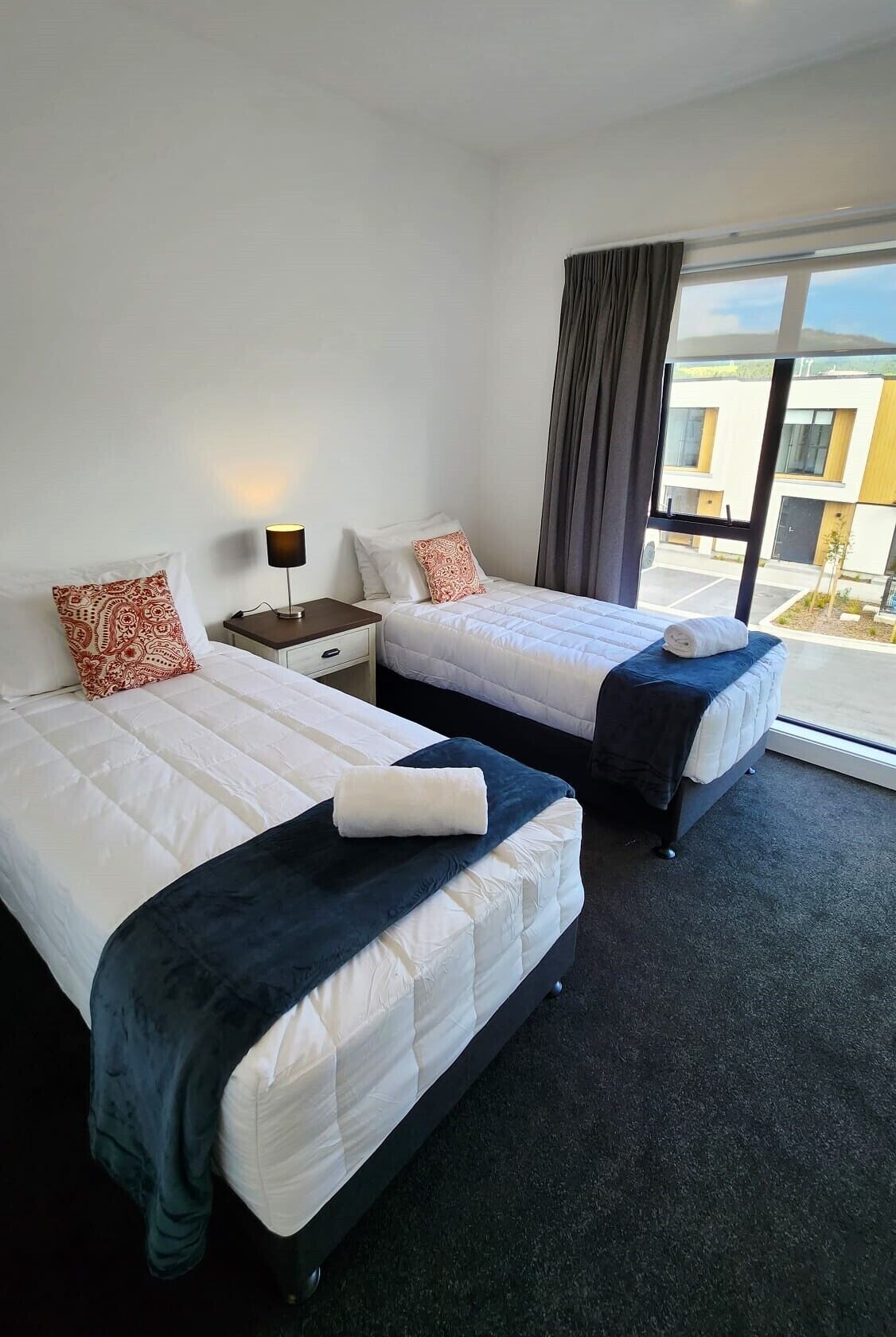 A hotel room with two beds, a nightstand with a lamp, and a large window offering a view of the building outside.