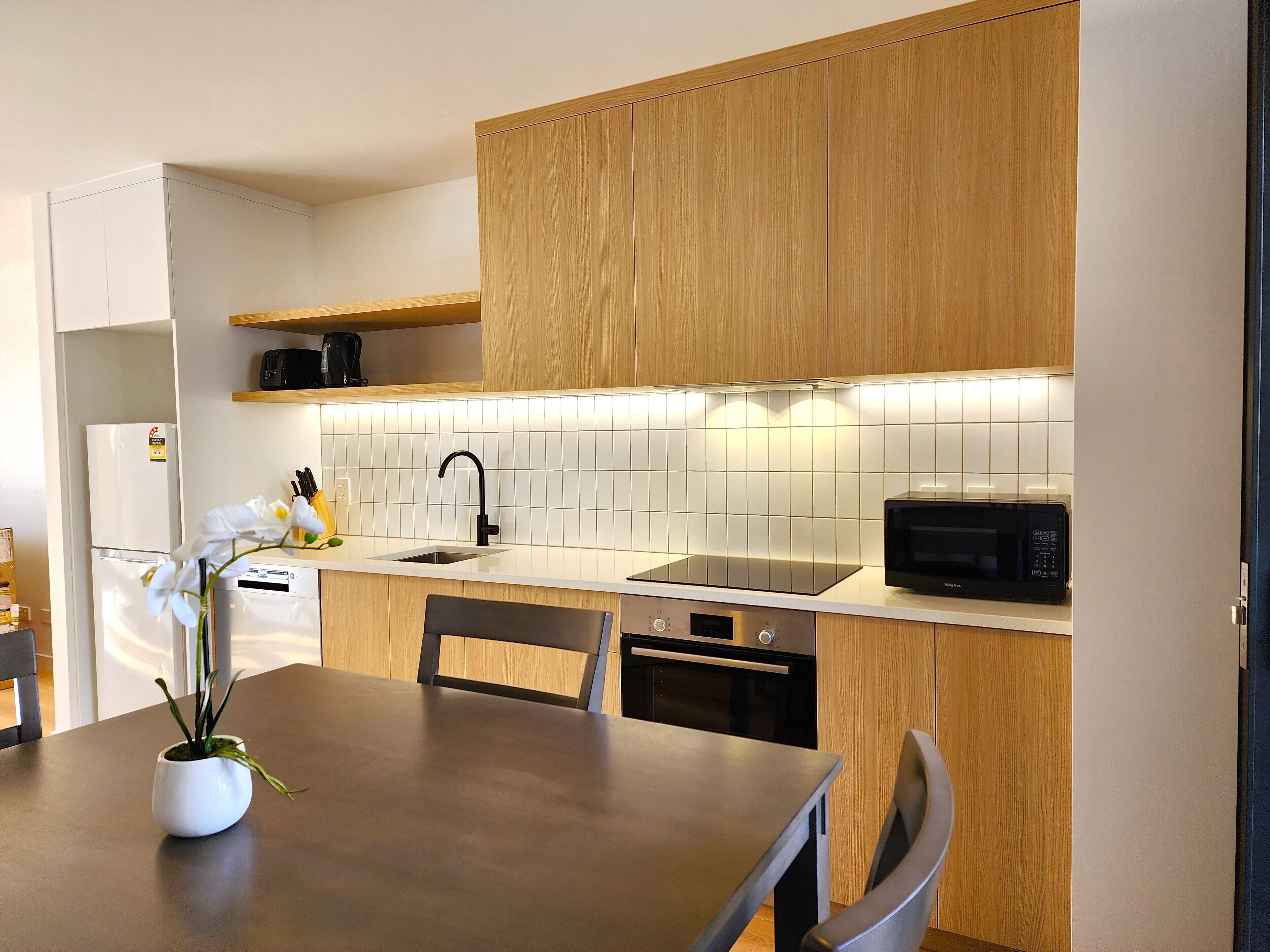 Une cuisine moderne avec des meubles en bois, un plan de travail blanc, un évier, un four à micro-ondes et un coin repas comprenant une table et des chaises.