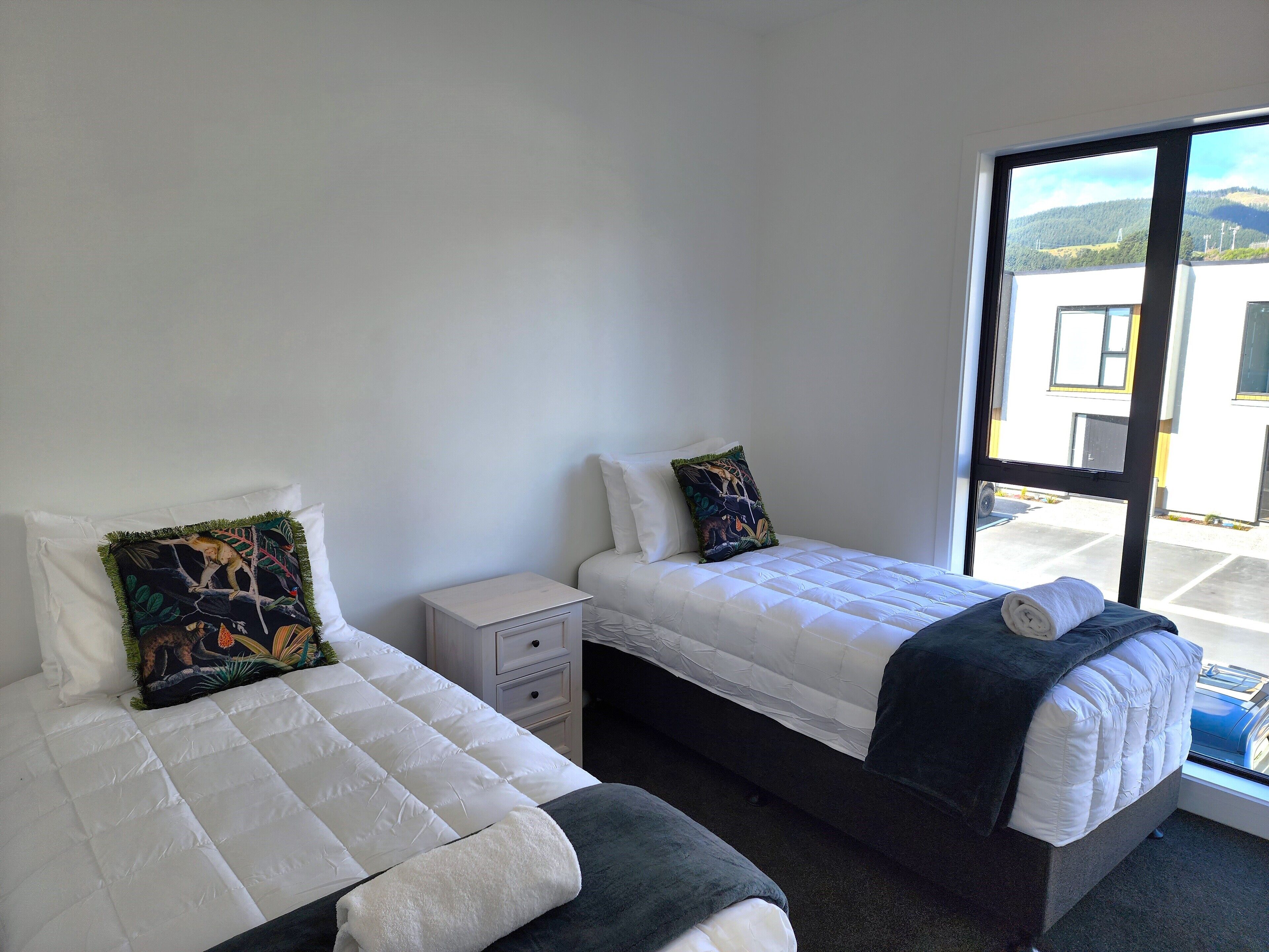 A room with two beds, a small bedside table, and a large window offering a view of buildings and hills.