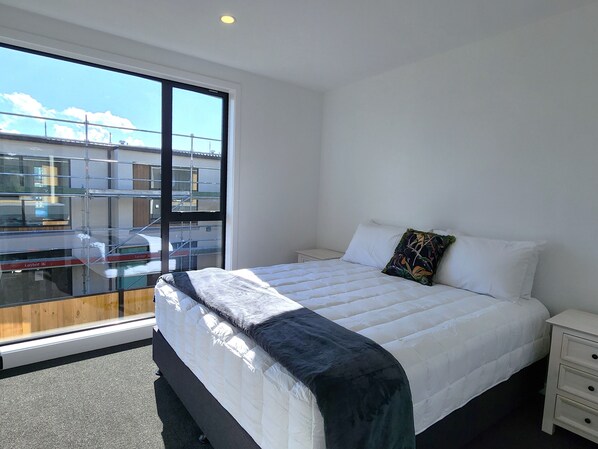 A modern bedroom with a large bed, a nightstand, and a window with a view of buildings. - U Suites on Trieste Way (Paraparaumu)