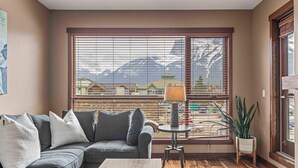 A living room with a sofa, a coffee table, a lamp, and a view of mountains through large windows. - Best Views of Canmore - 2Bed 2Bath (Canmore)