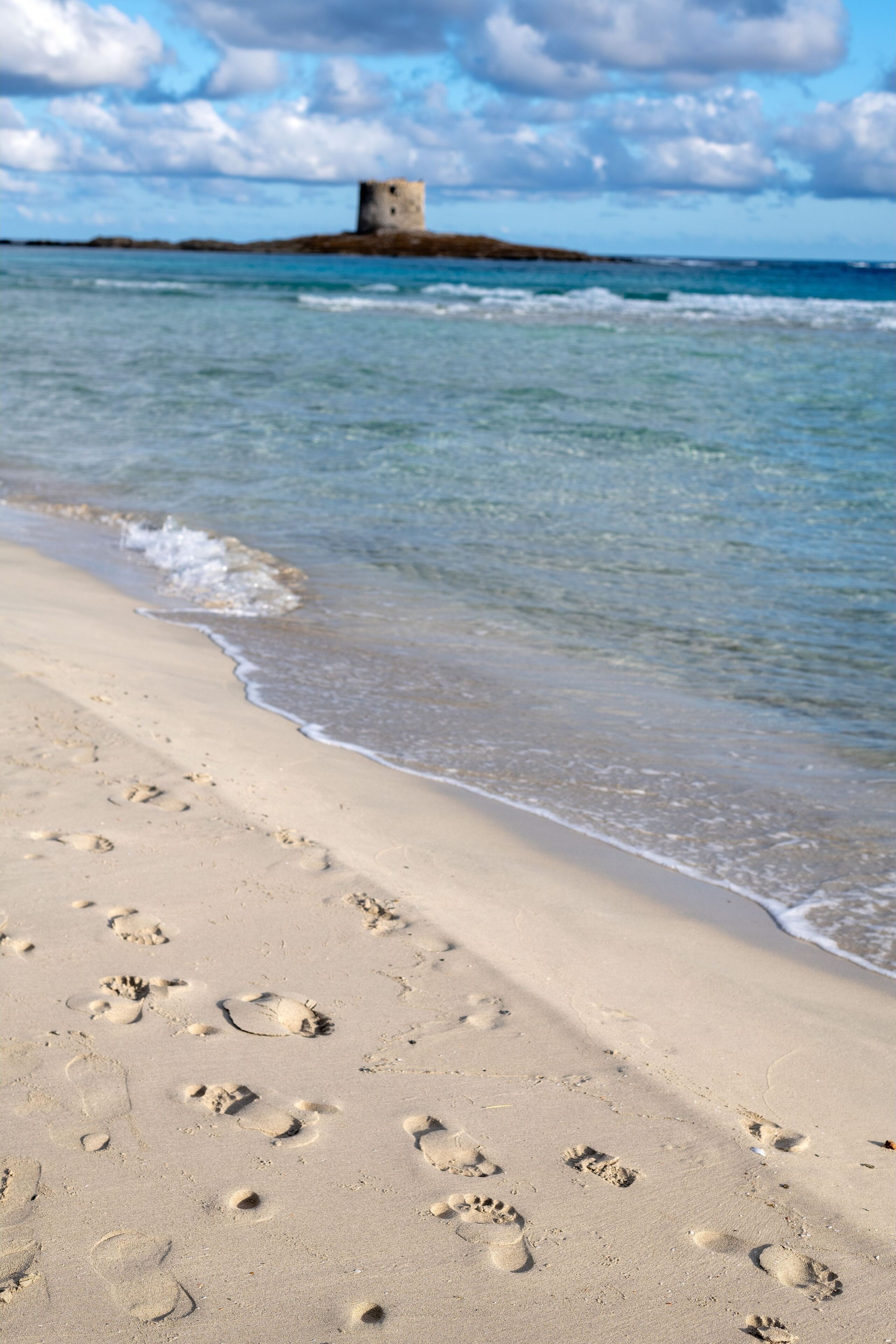 A sandy beach with footprints leading to the water and a tower in the distance.