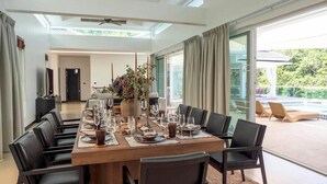 A dining area with a long table set for a meal, surrounded by chairs, with a view of a pool and outdoor seating. - Luxury Matahari Seaview (Kamala)