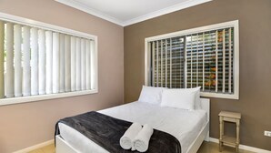 A bedroom with a bed, white pillows, a dark throw blanket, a small bedside table, and a window with blinds. - The Gallipoli Residence (Long Jetty)