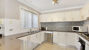 A modern kitchen with white cabinets, a tiled backsplash, stainless steel appliances, and a window with curtains. - The Gallipoli Residence (Long Jetty)
