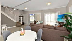 A living room with a sofa, a dining table with chairs, a TV, and a staircase. - The Gallipoli Residence (Long Jetty)
