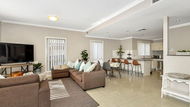 A modern living room with a sectional sofa, a flat-screen TV, and a dining area with bar stools. - The Gallipoli Residence (Long Jetty)