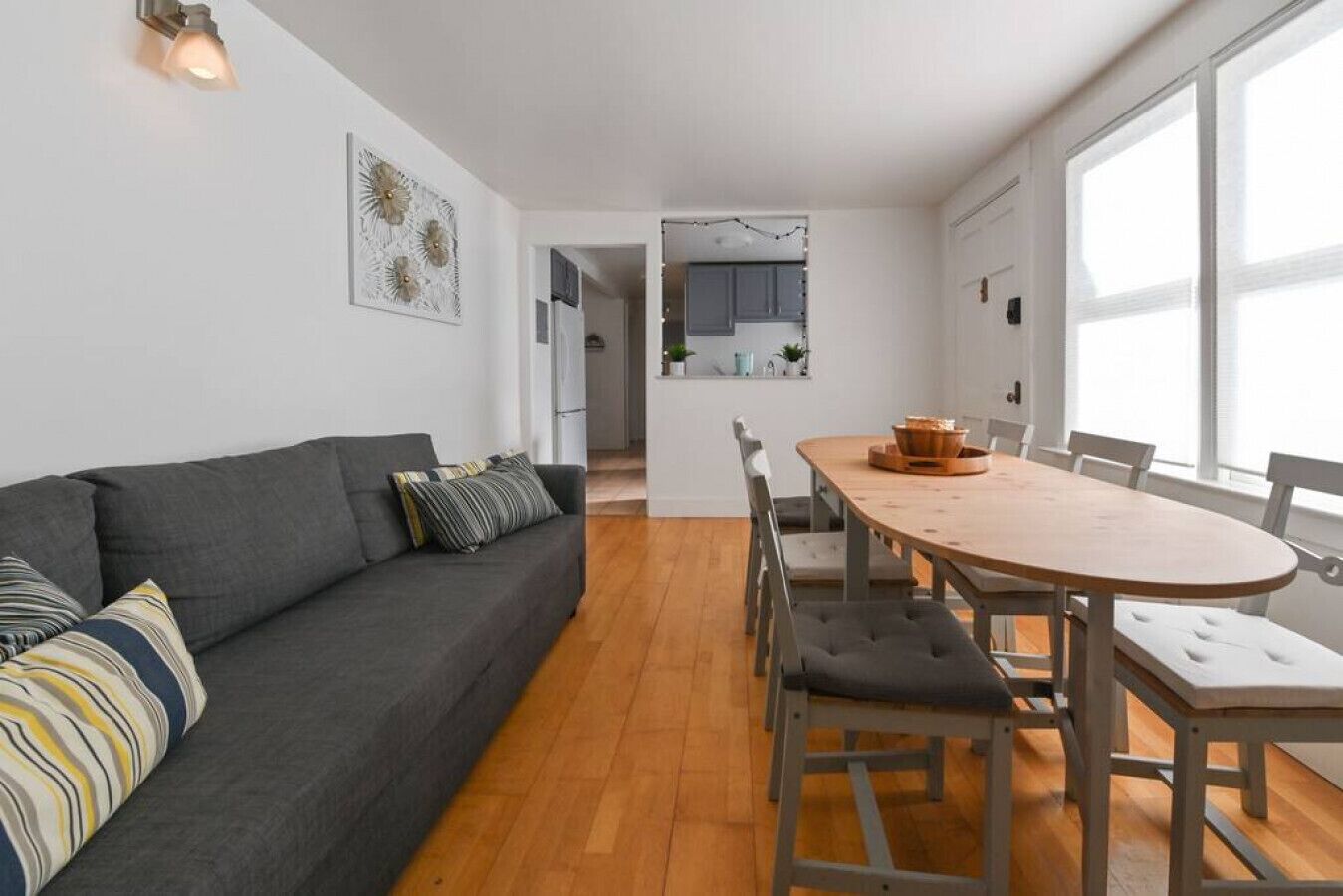 A modern living room with a grey sofa, a wooden dining table with chairs, and a kitchen in the background.