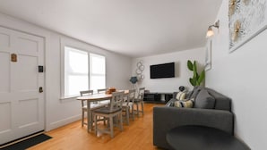 Una sala moderna con un sofá gris, una mesa de comedor con sillas, un televisor de pantalla plana en la pared y un gran ventanal que proporciona luz natural.