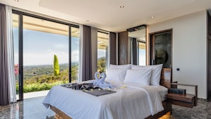 A modern bedroom with a large bed, a view of the landscape, and a wooden nightstand. - Villa Lovina Hill Paradise (Banjar)