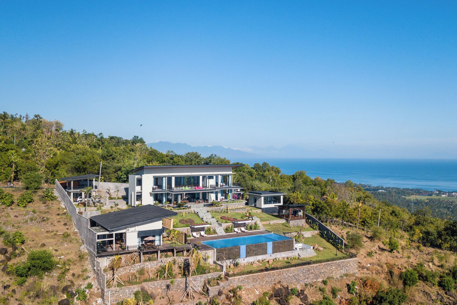 A modern two-story house with a large pool, surrounded by lush greenery and overlooking a coastal view.