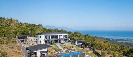 A modern two-story house with a large pool, surrounded by lush greenery and overlooking a coastal view.