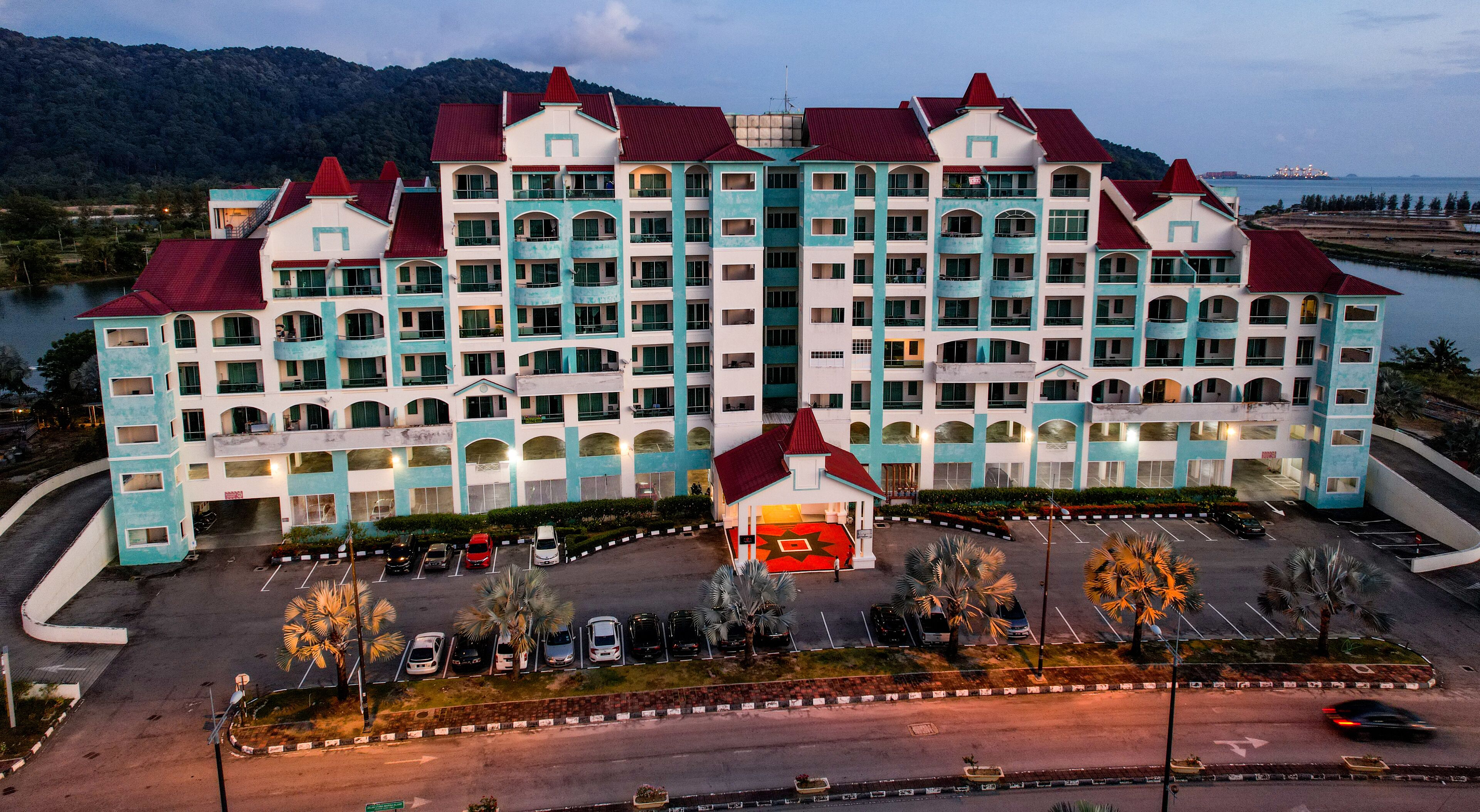 A large hotel with multiple floors and red roofs, surrounded by a parking area and palm trees.
