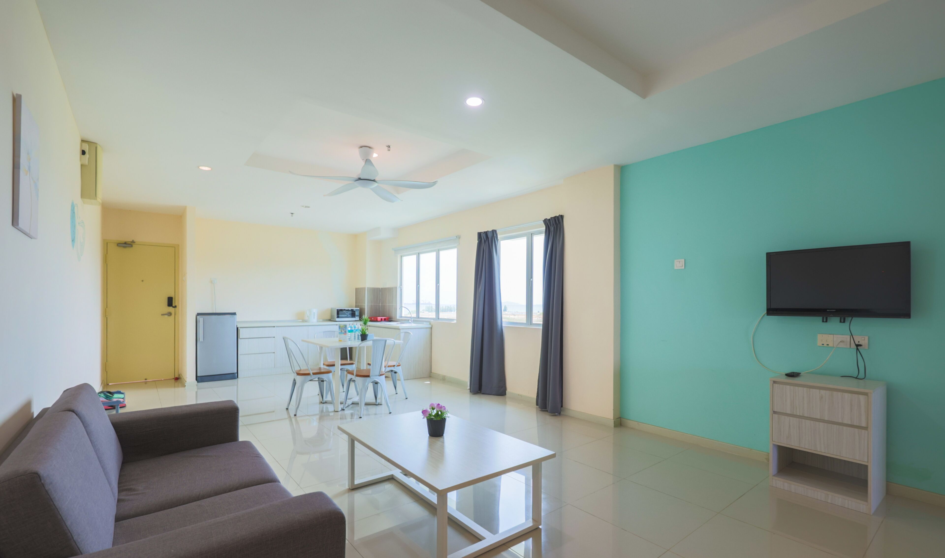 A modern living room with a sofa, a coffee table, and a TV stand. There is a kitchen area with a fridge, microwave, and sink.