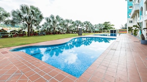 A swimming pool with tiled edges, surrounded by a tiled walkway, palm trees, and a building with balconies.