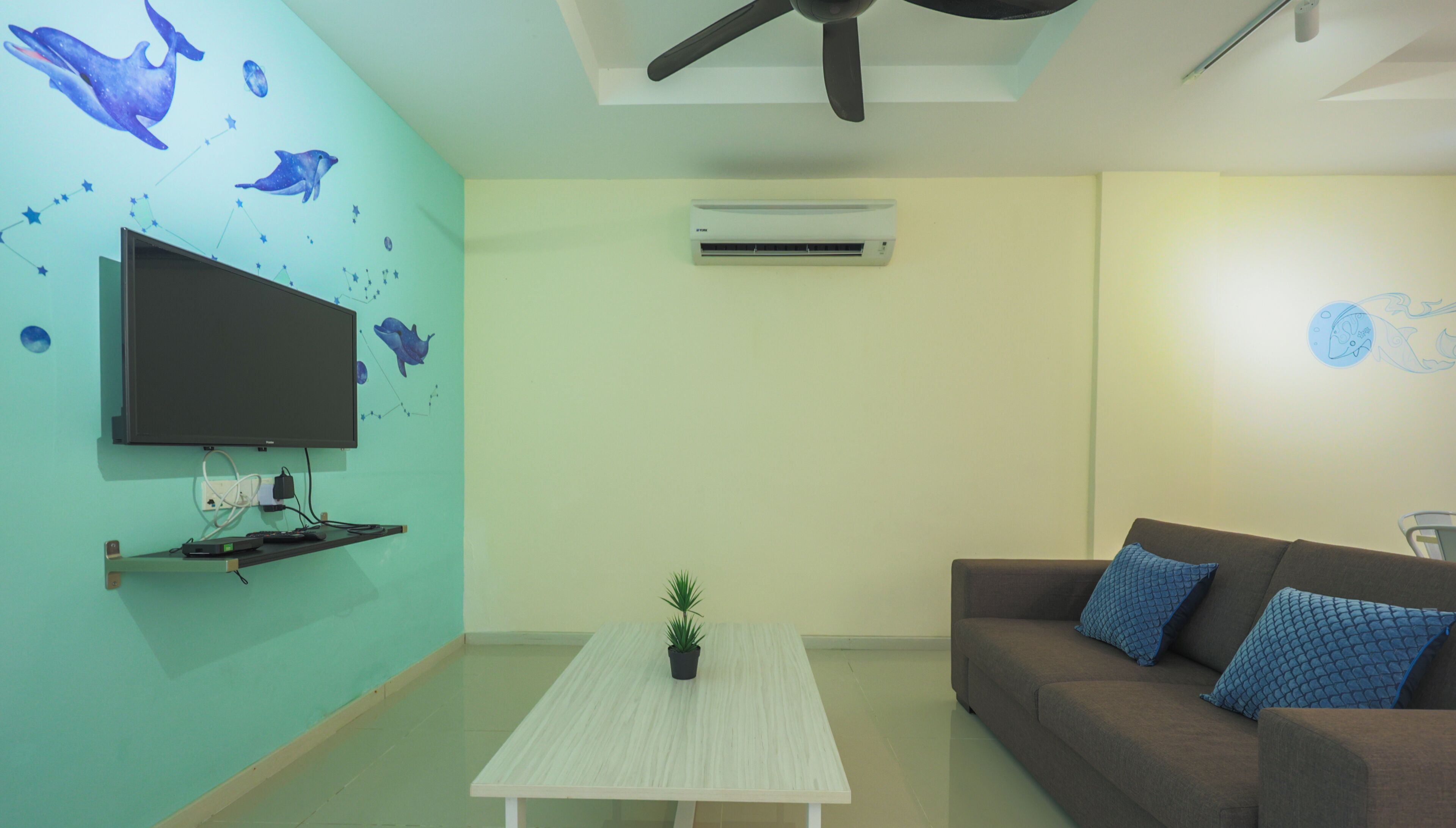 Ein Wohnzimmer mit Flachbildfernseher, Deckenventilator, Klimaanlage, Couchtisch und einer Couch mit gemusterten Kissen. An der Wand befindet sich ein Wandgemälde von einem Delfin.