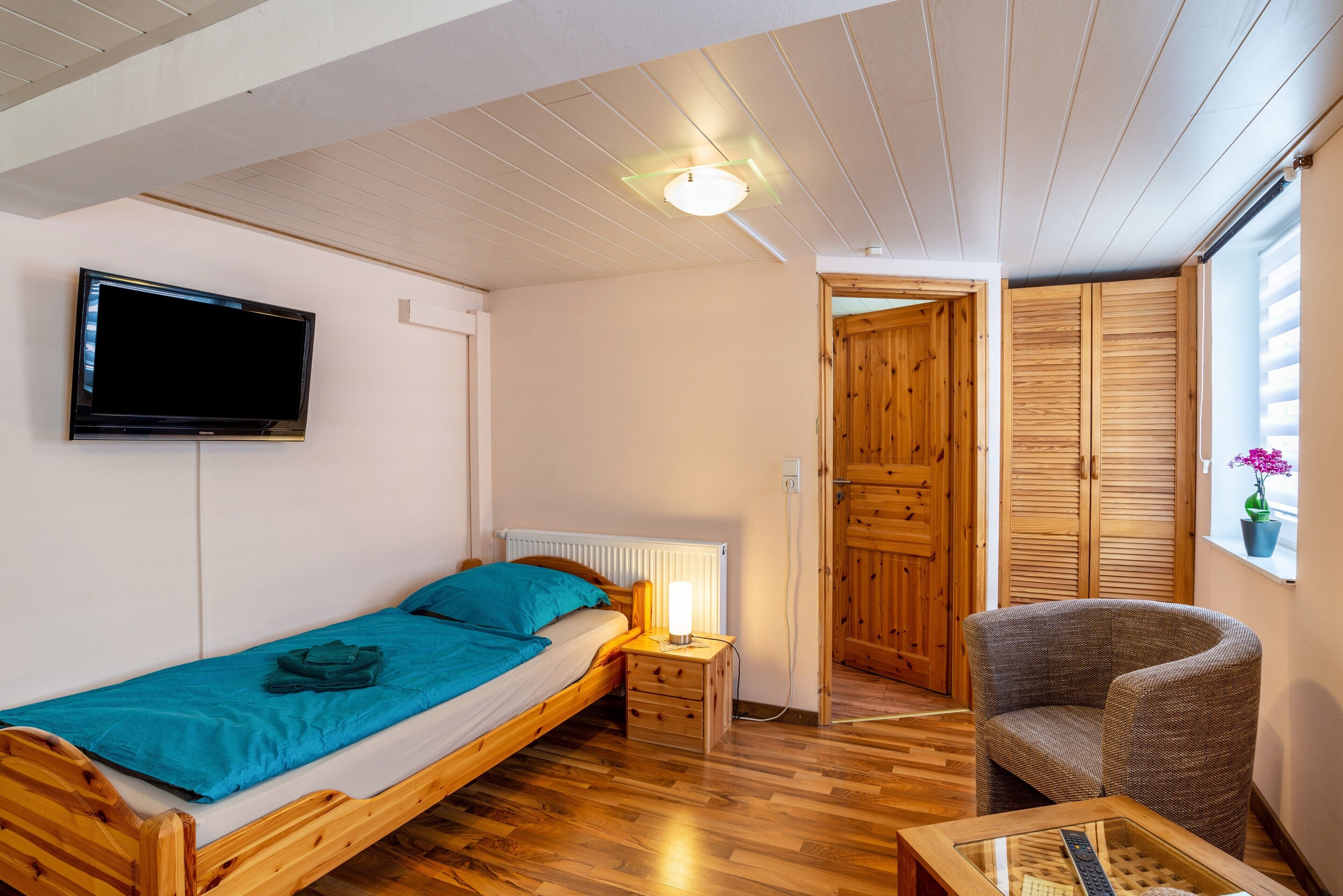 Ein kleines, gemütliches Zimmer mit einem Einzelbett, einem Sessel, einem Tisch, einem Fernseher und Holztüren.