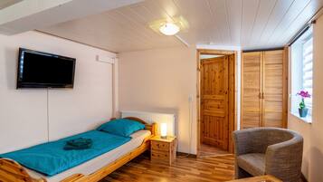 Ein kleines, gemütliches Zimmer mit einem Einzelbett, einem Sessel, einem Tisch, einem Fernseher und Holztüren.