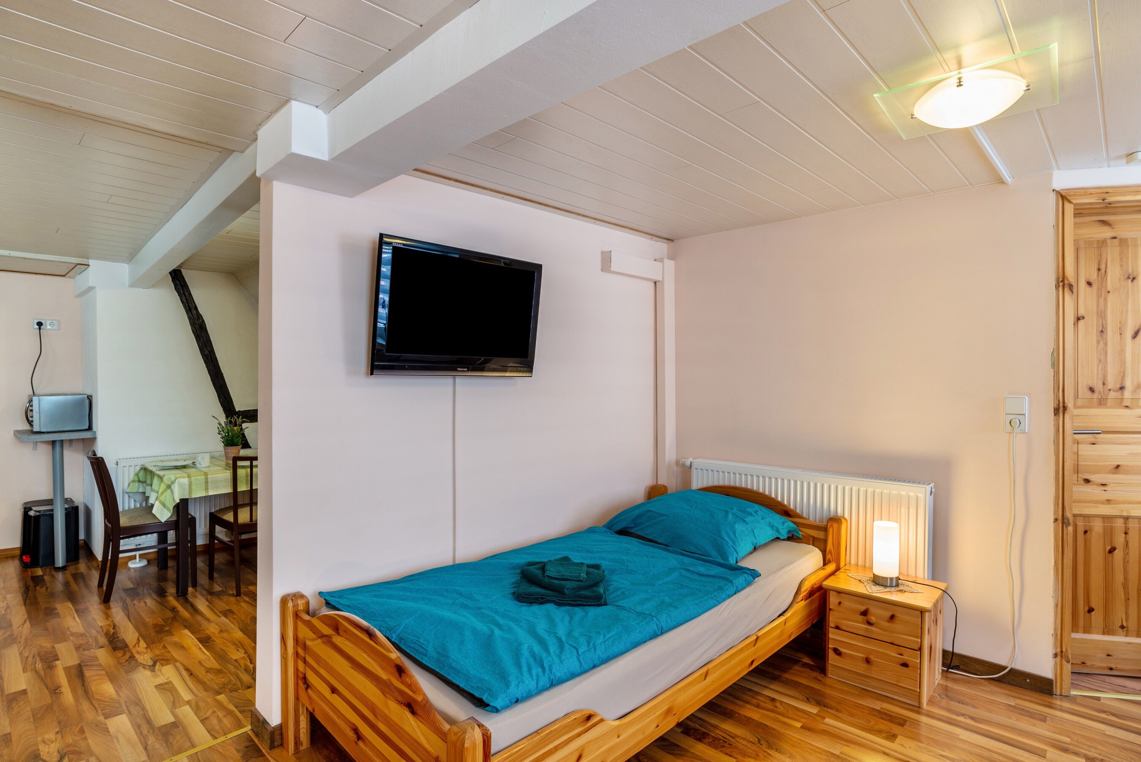 A single bed with a wooden frame, a bedside table with a lamp, a TV mounted on the wall, and a radiator.