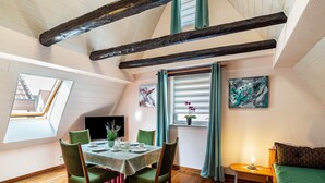 Un comedor con una mesa dispuesta para cuatro personas, una ventana con cortinas y un cuadro en la pared, y una cama con colcha verde.