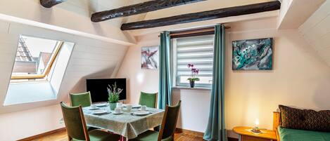 A dining area with a table set for four, a window with curtains, a painting on the wall, and a bed with a green bedspread.