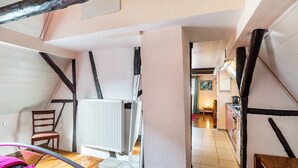 Una habitación con techo inclinado, vigas de madera, un radiador, una silla, una cama y vistas a otra habitación con zona de cocina.