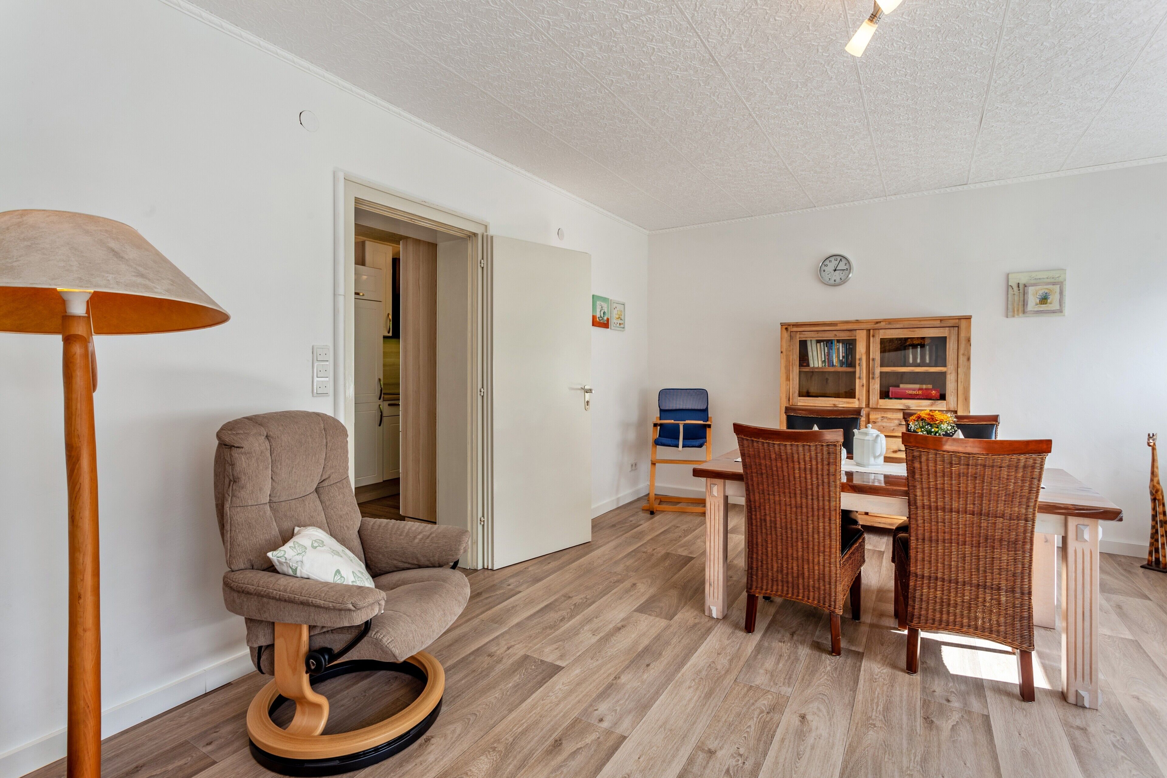 A room with a wooden floor, a round wooden chair, a dining table with wicker chairs, a bookshelf, and a lamp.