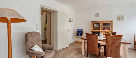 A room with a wooden floor, a round wooden chair, a dining table with wicker chairs, a bookshelf, and a lamp.