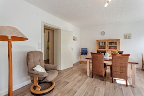A room with a wooden floor, a round wooden chair, a dining table with wicker chairs, a bookshelf, and a lamp.