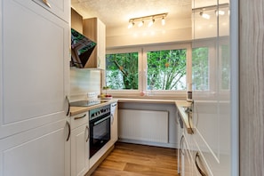 A modern kitchen with white cabinetry, a built-in oven, a window with a view of greenery, and a countertop with a kettle and fruit bowl.