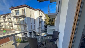 Un balcon avec deux fauteuils en osier et une table en verre, donnant sur une rue bordée d’immeubles.