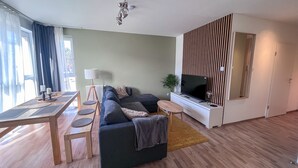 Un salon moderne comprenant un canapé d’angle, un téléviseur à écran plat, une table à manger avec des chaises et une grande fenêtre agrémentée de rideaux.