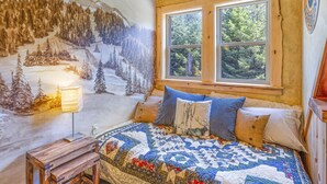 Un dormitorio acogedor con una cama grande, una ventana con vistas a árboles, un mural en la pared de un bosque nevado y una pequeña mesa de madera con una lámpara.