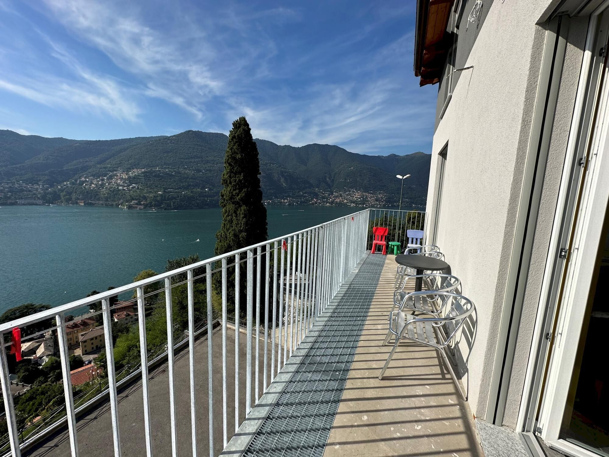 A balcony with a metal railing overlooking a lake, a mountain in the background, and a row of colorful chairs and tables.