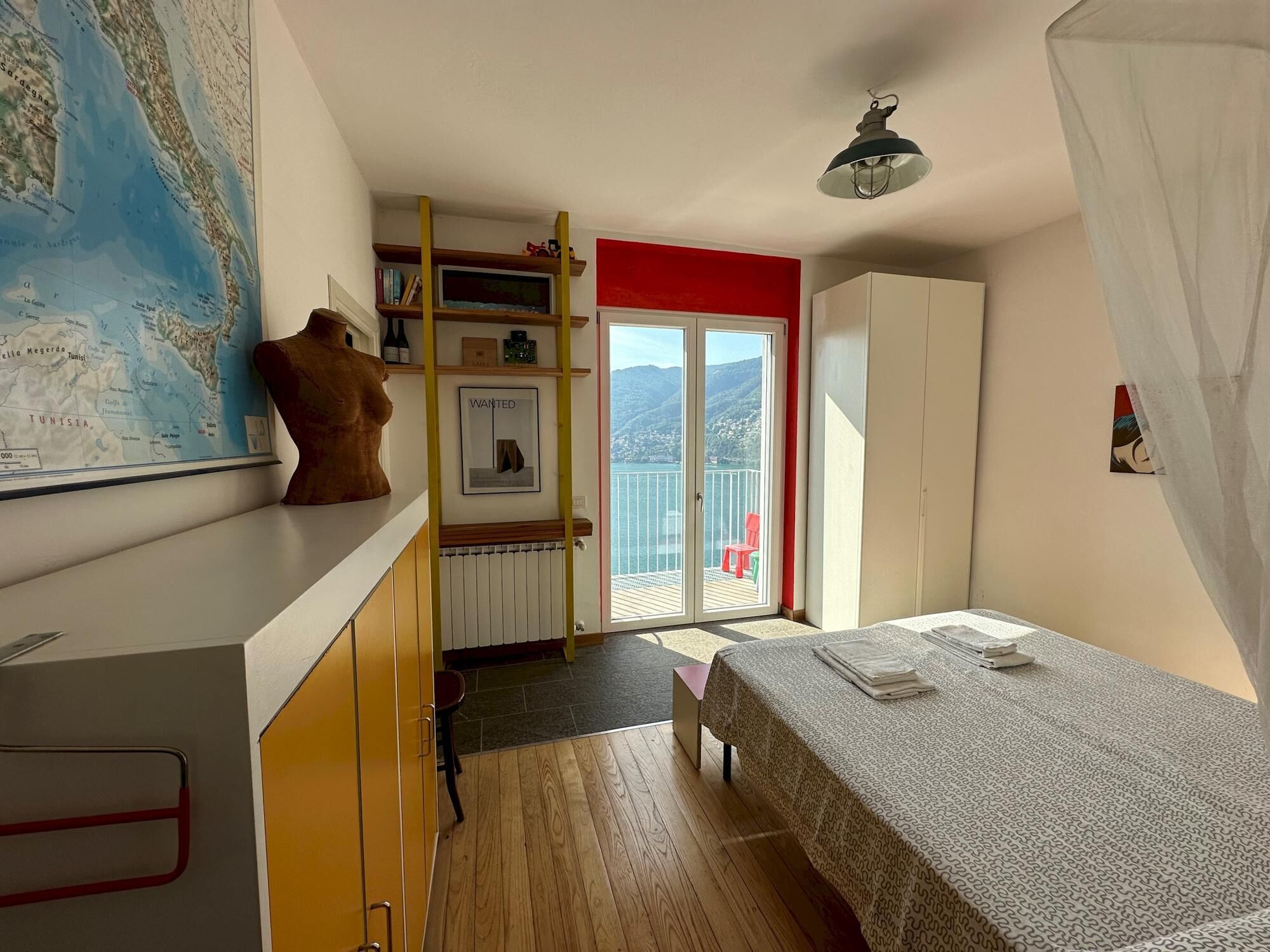 A bedroom with a bed, a map on the wall, a wardrobe, and a balcony with a view of the sea.