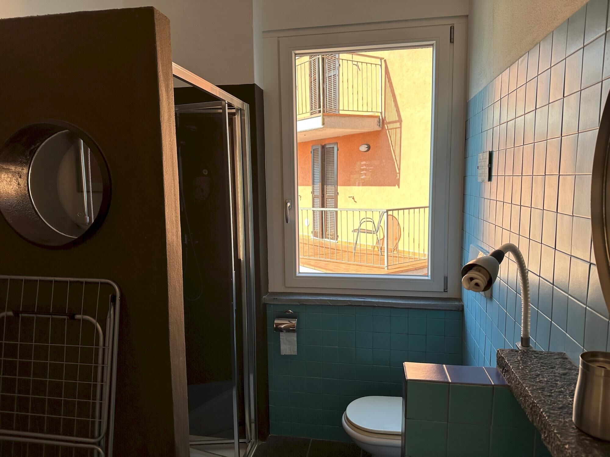 Egy fürdőszoba, amelyben WC, mosdókagyló, zuhanyzó és egy ablak található, melyről a balkonra nyílik kilátás.