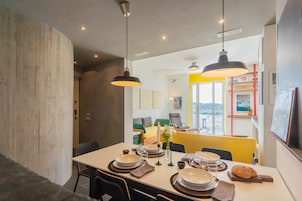 Un comedor con una mesa dispuesta para cuatro personas, vista a la ciudad a través de una ventana y una silla amarilla.