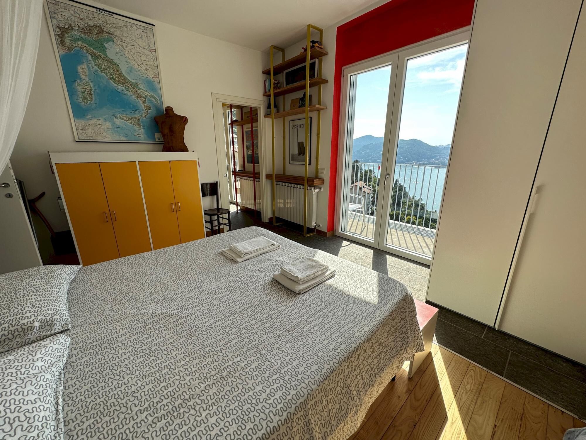 Un dormitorio con cama, armario, mapa en la pared, balcón con vista al mar y una pared de acento roja.