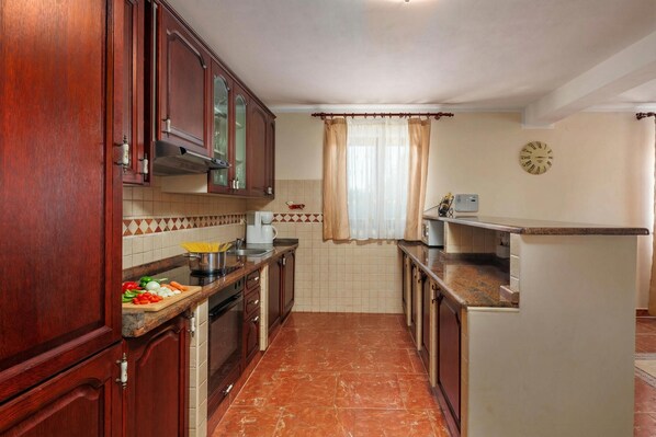 Una cocina con armarios de madera, salpicadero de azulejos, ventana con cortinas y un reloj en la pared.