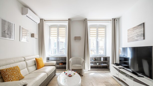 A modern living room with a sofa, ottoman, TV, and a view through large windows. - Le Meynadier dans le centre historique (Cannes)