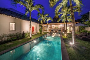 A swimming pool with a thatched roof structure, palm trees, and a well-lit outdoor area with seating. - Villa Palm Tree (Seminyak)