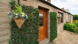 A stone house with a wooden door, a window with a basket of flowers, and a wall covered in climbing plants. - Heatherwood Cottage - Central Scone (Scone)