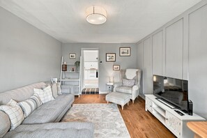 Un salon moderne comprenant un canapé, un fauteuil, un pouf, un téléviseur et un petit coin cuisine.