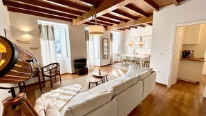 A spacious living area with a wooden staircase, a sofa, a dining table with chairs, and a kitchen area visible through an open door.